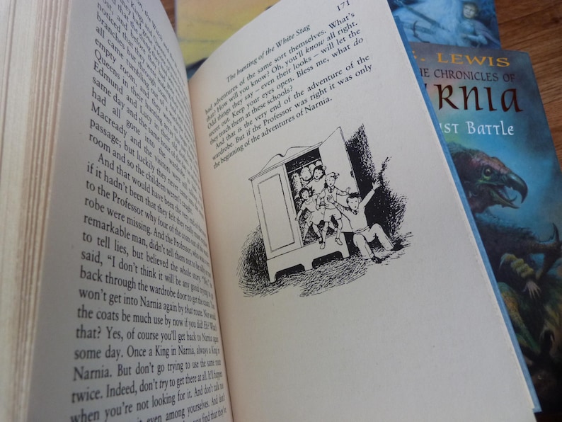 May include: A close-up of a book page with black and white illustrations of children entering a wardrobe. The text on the page reads "The hunting of the White Stag" and "Bless me, what do you know? Oh, you'll know all right. Children will say their eyes open... But if the Professor was right it was only the beginning of the adventures of Narnia."