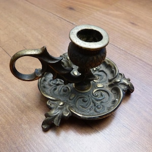 Peut inclure: Un bougeoir de type binard en laiton dot&eacute; d'une base orn&eacute;e et gaufr&eacute;e avec des volutes complexes et des motifs floraux. L'objet comprend une poign&eacute;e circulaire fix&eacute;e sur le c&ocirc;t&eacute; et une coupelle centrale con&ccedil;ue pour une bougie effil&eacute;e.