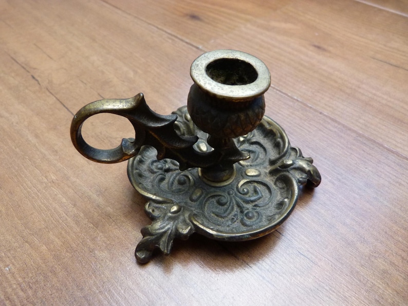 Peut inclure: Un bougeoir de type binard en laiton dot&eacute; d'une base orn&eacute;e et gaufr&eacute;e avec des volutes complexes et des motifs floraux. L'objet comprend une poign&eacute;e circulaire fix&eacute;e sur le c&ocirc;t&eacute; et une coupelle centrale con&ccedil;ue pour une bougie effil&eacute;e.