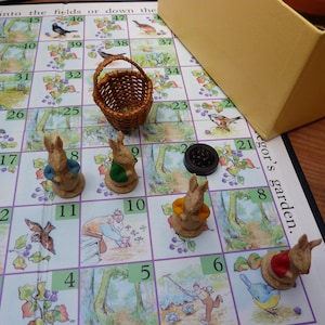 Könnte beinhalten: Ein Brettspiel mit einer Gartenillustration und nummerierten Feldern. Das Spiel enthält einen kleinen Weidenkorb, einen schwarzen Spielstein und Hasenfiguren in verschiedenen Outfits. Im Hintergrund befindet sich eine gelbe Box.