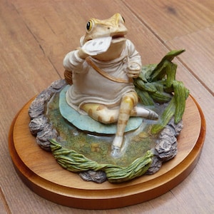 Peut inclure: Figurine fantaisiste d'une grenouille assise sur un nénuphar, tenant un objet blanc. La grenouille porte une tenue crème avec des jambes à motifs beige et blanc. La base est un cercle en bois avec une scène d'étang détaillée.