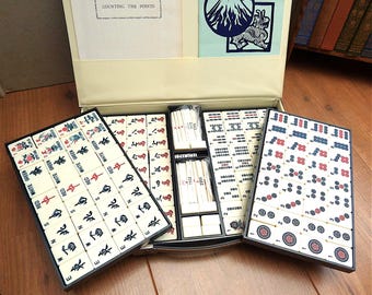 May include: A Mahjong game set in a cream-colored carrying case with two removable trays containing ivory-colored tiles. The tiles feature black, red, and blue ink designs, including characters, symbols, and numbers. Includes a manual titled Counting the Points.