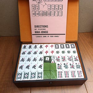 May include: A Mah-Jongg game set in a dark brown case with a light orange interior. The set includes white tiles with black, red, and green symbols, two small white dice, and a pink instruction booklet titled DIRECTIONS OF PLAYING MAH-JONGG.