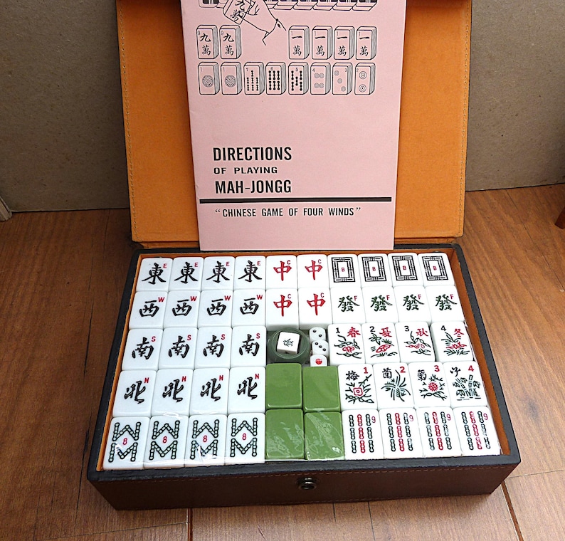 May include: A Mah-Jongg game set in a dark brown case with a light orange interior. The set includes white tiles with black, red, and green symbols, two small white dice, and a pink instruction booklet titled DIRECTIONS OF PLAYING MAH-JONGG.