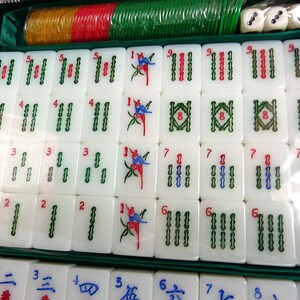 May include: A Mahjong set featuring white tiles with green, red, and blue engraved symbols, including bamboo patterns and bird illustrations. The set is organized in a green case with colorful plastic chips and three white dice included at the top.