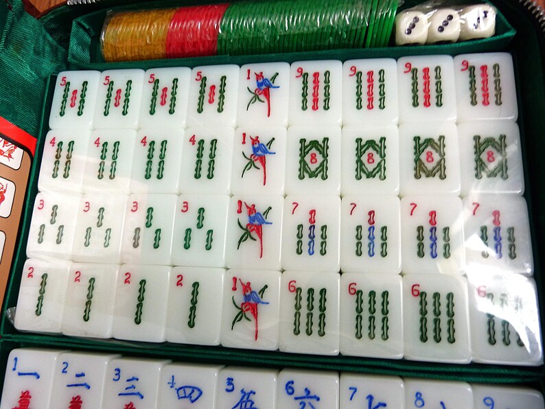 May include: A Mahjong set featuring white tiles with green, red, and blue engraved symbols, including bamboo patterns and bird illustrations. The set is organized in a green case with colorful plastic chips and three white dice included at the top.