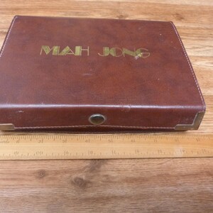 May include: A rectangular brown Mah Jong case with gold lettering on the lid, featuring metal corner reinforcements and a central snap closure. A wooden ruler is placed in front, showing the case is approximately 13 inches in length.