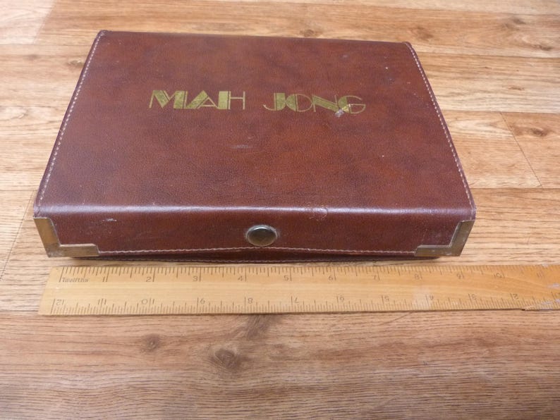 May include: A rectangular brown Mah Jong case with gold lettering on the lid, featuring metal corner reinforcements and a central snap closure. A wooden ruler is placed in front, showing the case is approximately 13 inches in length.