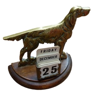 May include: A vintage brass dog figurine with a calendar displaying "FRIDAY DECEMBER 25". The dog is standing on a wooden base. The figurine is a decorative item.