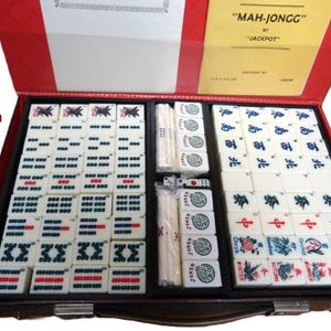 Peut inclure: Un jeu de Mah-Jongg de la marque Jackpot dans une mallette rouge. Le set comprend 152 tuiles avec divers symboles, caract&egrave;res et motifs floraux, ainsi que des b&acirc;tonnets de pari blancs et des d&eacute;s. Le couvercle int&eacute;rieur porte une &eacute;tiquette jaune.