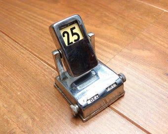 May include: A chrome-finished perpetual desk calendar with a rectangular pivoting head displaying the number 25. The base features two side-mounted knurled knobs and horizontal windows showing the text DEC and FRI in black lettering.