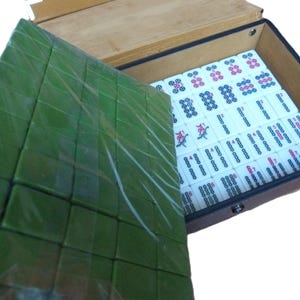 May include: A mahjong set housed in a brown rectangular case with a hinged lid. The interior contains white tiles with black, red, and blue markings. A separate tray of green tiles is positioned on top, partially covering the main set of tiles.