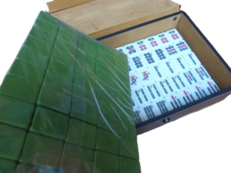 May include: A mahjong set housed in a brown rectangular case with a hinged lid. The interior contains white tiles with black, red, and blue markings. A separate tray of green tiles is positioned on top, partially covering the main set of tiles.