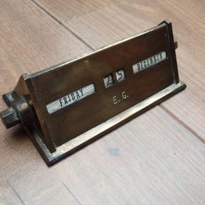 May include: A vintage brass perpetual calendar showing "25 DECEMBER" and "FRIDAY". The calendar has a rectangular base and a rotating mechanism. The initials "E. G." are visible. It is set on a wooden surface, a classic desk accessory.
