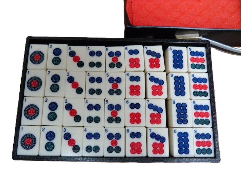 Peut inclure: Un plateau rectangulaire noir contenant un ensemble de tuiles de mah-jong blanc cass&eacute; dispos&eacute;es en quatre rang&eacute;es. Chaque tuile pr&eacute;sente un chiffre de un &agrave; neuf, repr&eacute;sent&eacute; par des motifs circulaires rouges, bleus et verts. Les tuiles sont lisses.