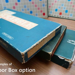 Puede incluir: Primer plano de tres cajas de juego azules y verdes apiladas una encima de la otra. La caja superior tiene la palabra "SCRABBLE" impresa en blanco en la parte delantera. Las cajas est&aacute;n desgastadas y muestran signos de edad.