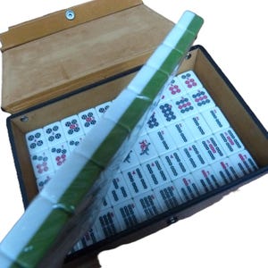 May include: A mahjong set featuring white tiles with green backs, arranged in a rectangular case with a tan lid. The tiles display various black, red, and blue character markings and numerical symbols, with a row of tiles held upright in the foreground.