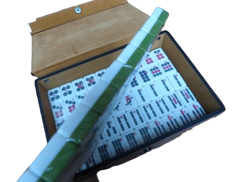 May include: A mahjong set featuring white tiles with green backs, arranged in a rectangular case with a tan lid. The tiles display various black, red, and blue character markings and numerical symbols, with a row of tiles held upright in the foreground.