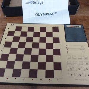May include: A black electronic chessboard with a brown and beige chessboard design. The board has a digital display with buttons for game settings and piece selection. The text "SciSys OLYMPIADE" is printed on the board.