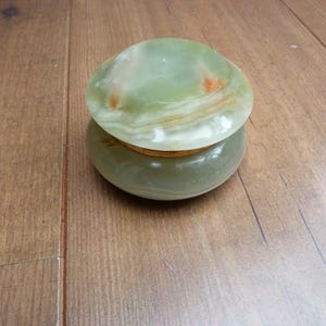May include: A round, polished green onyx stone trinket box with a smooth, domed lid. The stone features natural banding in shades of pale green, white, and translucent cream, with small, distinct orange-brown inclusions visible on the lid surface.