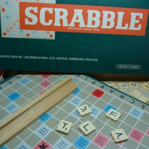 Puede incluir: Un primer plano de un tablero de juego de Scrabble con fichas colocadas en el tablero. Las fichas deletrean las palabras "SCRABBLE" y "TILT". El tablero de juego est&aacute; hecho de una cuadr&iacute;cula azul claro y blanca con cuadrados rojos y azules. Las fichas son blancas con letras negras. El tablero de juego est&aacute; rodeado por un marco de madera.
