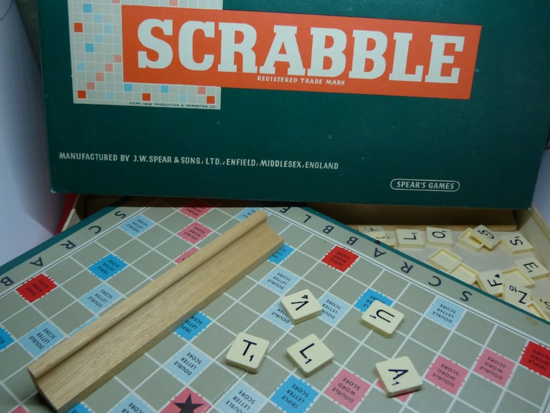 Puede incluir: Un primer plano de un tablero de juego de Scrabble con fichas colocadas en el tablero. Las fichas deletrean las palabras "SCRABBLE" y "TILT". El tablero de juego est&aacute; hecho de una cuadr&iacute;cula azul claro y blanca con cuadrados rojos y azules. Las fichas son blancas con letras negras. El tablero de juego est&aacute; rodeado por un marco de madera.