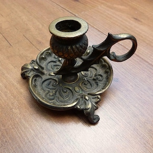 Peut inclure: Un bougeoir de type binard en laiton dot&eacute; d'une base circulaire orn&eacute;e de motifs &agrave; volutes en relief, d'une douille centrale au design textur&eacute; rappelant un ananas et d'une anse incurv&eacute;e fix&eacute;e sur le c&ocirc;t&eacute; de la coupelle &agrave; bougie.