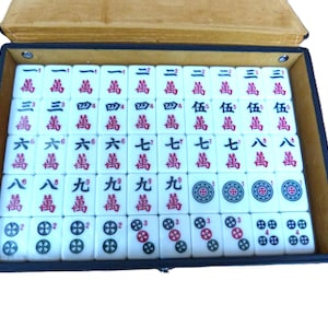 May include: A rectangular black storage case containing a set of white Mahjong tiles arranged in rows. The tiles feature red and black Chinese characters, Arabic numerals, and circular floral patterns. The case has a tan interior lining.