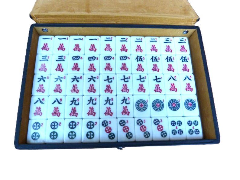 May include: A rectangular black storage case containing a set of white Mahjong tiles arranged in rows. The tiles feature red and black Chinese characters, Arabic numerals, and circular floral patterns. The case has a tan interior lining.
