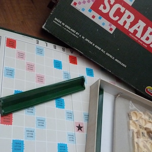 Puede incluir: Un primer plano de un tablero de juego de Scrabble con las fichas de letras y un portafichas verde. El tablero est&aacute; hecho de pl&aacute;stico blanco con una cuadr&iacute;cula de casillas. Las casillas est&aacute;n marcadas con diferentes valores de puntos y bonificaciones especiales. El juego est&aacute; hecho en Inglaterra por J.W. Spear & Sons PLC, Enfield, Middlesex.
