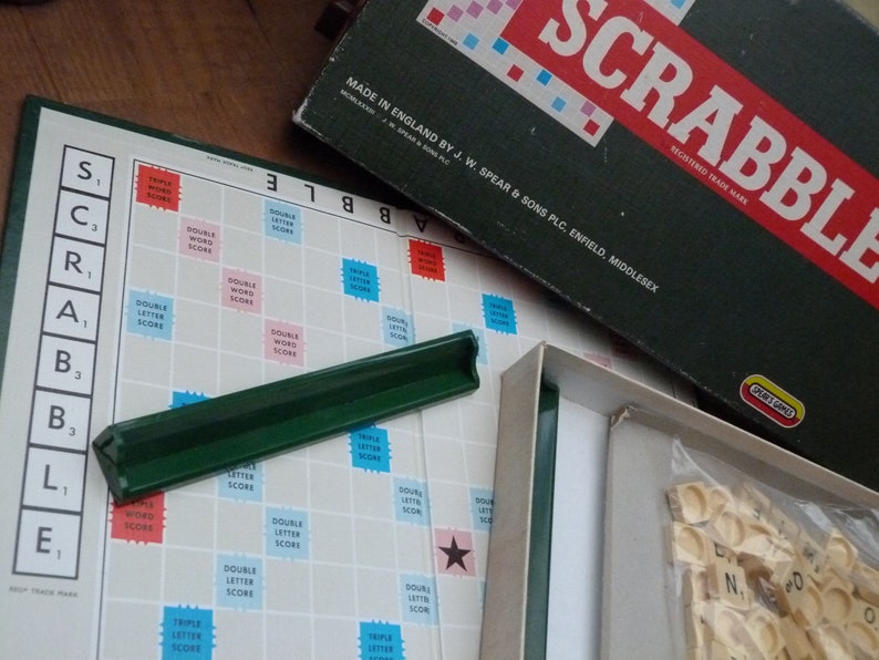 Puede incluir: Un primer plano de un tablero de juego de Scrabble con las fichas de letras y un portafichas verde. El tablero est&aacute; hecho de pl&aacute;stico blanco con una cuadr&iacute;cula de casillas. Las casillas est&aacute;n marcadas con diferentes valores de puntos y bonificaciones especiales. El juego est&aacute; hecho en Inglaterra por J.W. Spear & Sons PLC, Enfield, Middlesex.