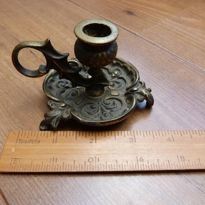 Peut inclure: Un bougeoir de type chamberstick en laiton avec une base orn&eacute;e en relief et une poign&eacute;e en boucle. Une r&egrave;gle en bois est plac&eacute;e devant le bougeoir, indiquant que le diam&egrave;tre de la base est d'environ 9 cm de large.