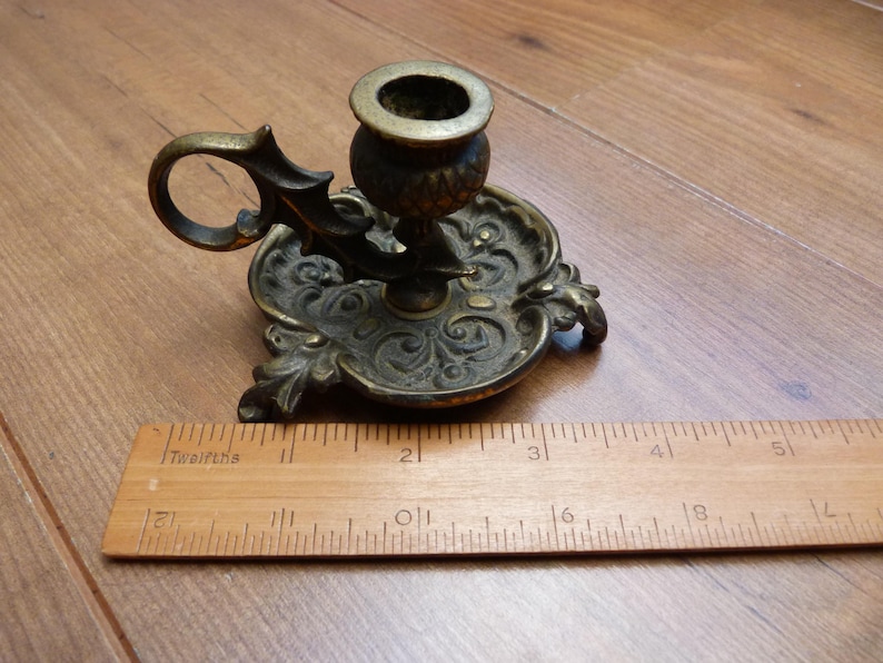 Peut inclure: Un bougeoir de type chamberstick en laiton avec une base orn&eacute;e en relief et une poign&eacute;e en boucle. Une r&egrave;gle en bois est plac&eacute;e devant le bougeoir, indiquant que le diam&egrave;tre de la base est d'environ 9 cm de large.