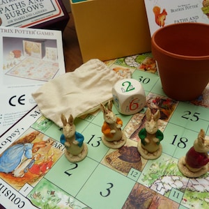 Könnte beinhalten: Ein Brettspiel mit Beatrix Potter-Thema, mit einem Spielbrett, Würfeln, Hasenfiguren und einem Terrakotta-Topf. Das Spielbrett hat ein Rastermuster mit Illustrationen und Zahlen. Das Spiel heißt "Paths and Burrows".
