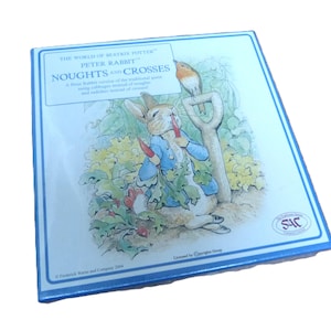 May include: A square Peter Rabbit game box with a blue border. The box features an illustration of Peter Rabbit holding carrots with a bird perched on a shovel. Text includes "Noughts and Crosses" and "The World of Beatrix Potter."