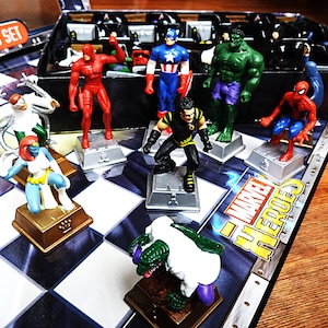 May include: A Marvel Heroes chess set featuring various character figurines on a black and white checkered board. The set includes figures of Daredevil, Captain America, Hulk, Spider-Man, Doctor Octopus, Mystique, and others on individual bases.