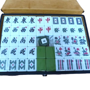 May include: A Mahjong set contained in a rectangular box with a tan lid. The set includes white tiles featuring black, red, green, and blue characters, symbols, and numbers. There are also plain green tiles, small white dice, and a circular token.