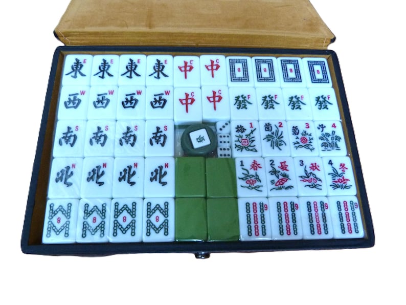 May include: A Mahjong set contained in a rectangular box with a tan lid. The set includes white tiles featuring black, red, green, and blue characters, symbols, and numbers. There are also plain green tiles, small white dice, and a circular token.