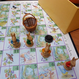 May include: A board game featuring a grid with numbered squares and illustrations of birds and garden scenes. Four small rabbit figurines in blue, green, orange, and red jackets are placed on the board alongside a miniature woven basket and a black token.