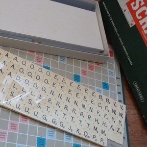 Puede incluir: Un primer plano de un tablero de juego de Scrabble con fichas en una bolsa de pl&aacute;stico. Las fichas est&aacute;n ordenadas alfab&eacute;ticamente e incluyen letras como A, E, I, O, U y otras. El tablero est&aacute; hecho de un material azul claro con un patr&oacute;n de cuadr&iacute;cula. El juego est&aacute; fabricado por J.W. Spear & Sons Ltd. en Enfield, Middlesex, Inglaterra.