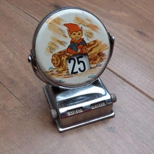 Peut inclure: Calendrier de bureau vintage en métal avec un cadran circulaire représentant un elfe de dessin animé sur une bûche. La date affichée est le "25". La base indique "DEC THU". Le calendrier est argenté et repose sur une surface en bois.