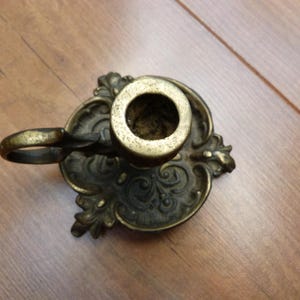 Peut inclure: Un bougeoir en laiton dot&eacute; d'une base orn&eacute;e et gaufr&eacute;e avec des motifs en volutes et un plateau circulaire. L'objet comprend une petite poign&eacute;e incurv&eacute;e fix&eacute;e sur le c&ocirc;t&eacute; et une douille cylindrique centrale con&ccedil;ue pour une bougie effil&eacute;e.