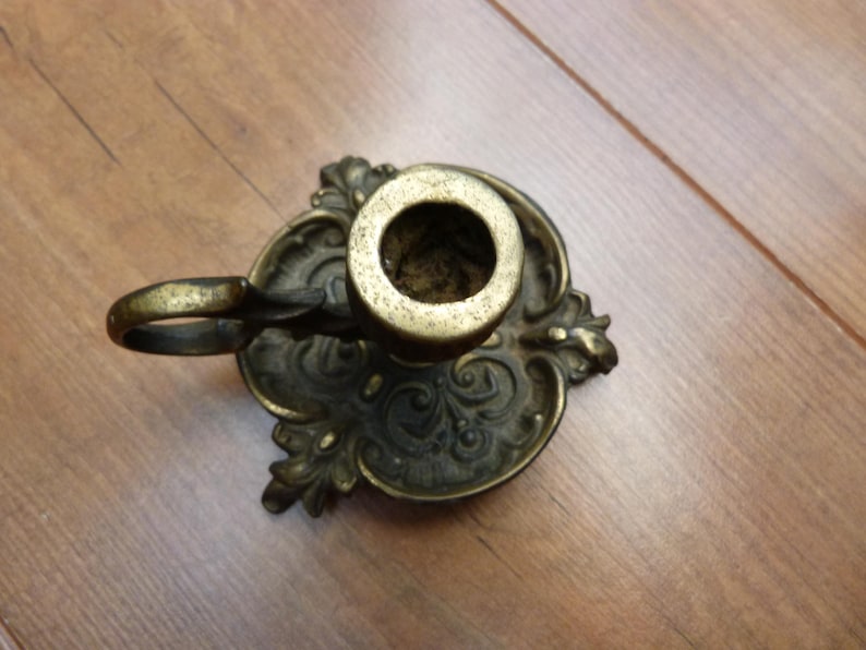Peut inclure: Un bougeoir en laiton dot&eacute; d'une base orn&eacute;e et gaufr&eacute;e avec des motifs en volutes et un plateau circulaire. L'objet comprend une petite poign&eacute;e incurv&eacute;e fix&eacute;e sur le c&ocirc;t&eacute; et une douille cylindrique centrale con&ccedil;ue pour une bougie effil&eacute;e.