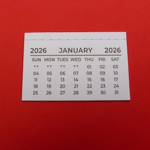 May include: A small, rectangular calendar for January 2026, printed on white paper. The calendar features the days of the week across the top, with the month and year clearly displayed. The background is a solid red.