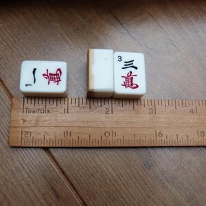 以下が含まれることがあります： 赤と黒の中国語の文字と数字の1と3が描かれた3枚の白い麻雀牌。木製の定規に沿って配置されており、各牌の幅は約1.9cm、高さは約2.5cmであることが示されています。
