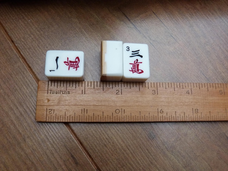 以下が含まれることがあります： 赤と黒の中国語の文字と数字の1と3が描かれた3枚の白い麻雀牌。木製の定規に沿って配置されており、各牌の幅は約1.9cm、高さは約2.5cmであることが示されています。