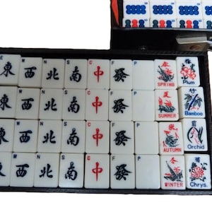 Peut inclure: Un ensemble de tuiles de Mahjong blanches dispos&eacute;es dans un plateau noir. Les tuiles pr&eacute;sentent des caract&egrave;res chinois noirs et rouges, des lettres directionnelles E, W, N, S, C, F, P, et des illustrations color&eacute;es &eacute;tiquet&eacute;es Spring, Summer, Autumn, Winter, Plum, Bamboo, Orchid et Chrys.