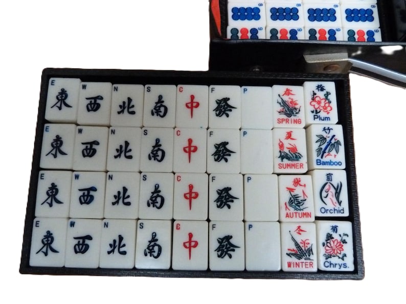 Peut inclure: Un ensemble de tuiles de Mahjong blanches dispos&eacute;es dans un plateau noir. Les tuiles pr&eacute;sentent des caract&egrave;res chinois noirs et rouges, des lettres directionnelles E, W, N, S, C, F, P, et des illustrations color&eacute;es &eacute;tiquet&eacute;es Spring, Summer, Autumn, Winter, Plum, Bamboo, Orchid et Chrys.