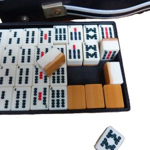 Peut inclure: Un ensemble de tuiles de Mahjong blanches et marron clair dispos&eacute;es dans un plateau rectangulaire noir. Les tuiles pr&eacute;sentent des marquages et symboles num&eacute;riques noirs et rouges. Une tuile est plac&eacute;e s&eacute;par&eacute;ment sous le plateau.
