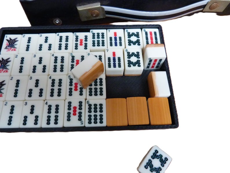 Peut inclure: Un ensemble de tuiles de Mahjong blanches et marron clair dispos&eacute;es dans un plateau rectangulaire noir. Les tuiles pr&eacute;sentent des marquages et symboles num&eacute;riques noirs et rouges. Une tuile est plac&eacute;e s&eacute;par&eacute;ment sous le plateau.